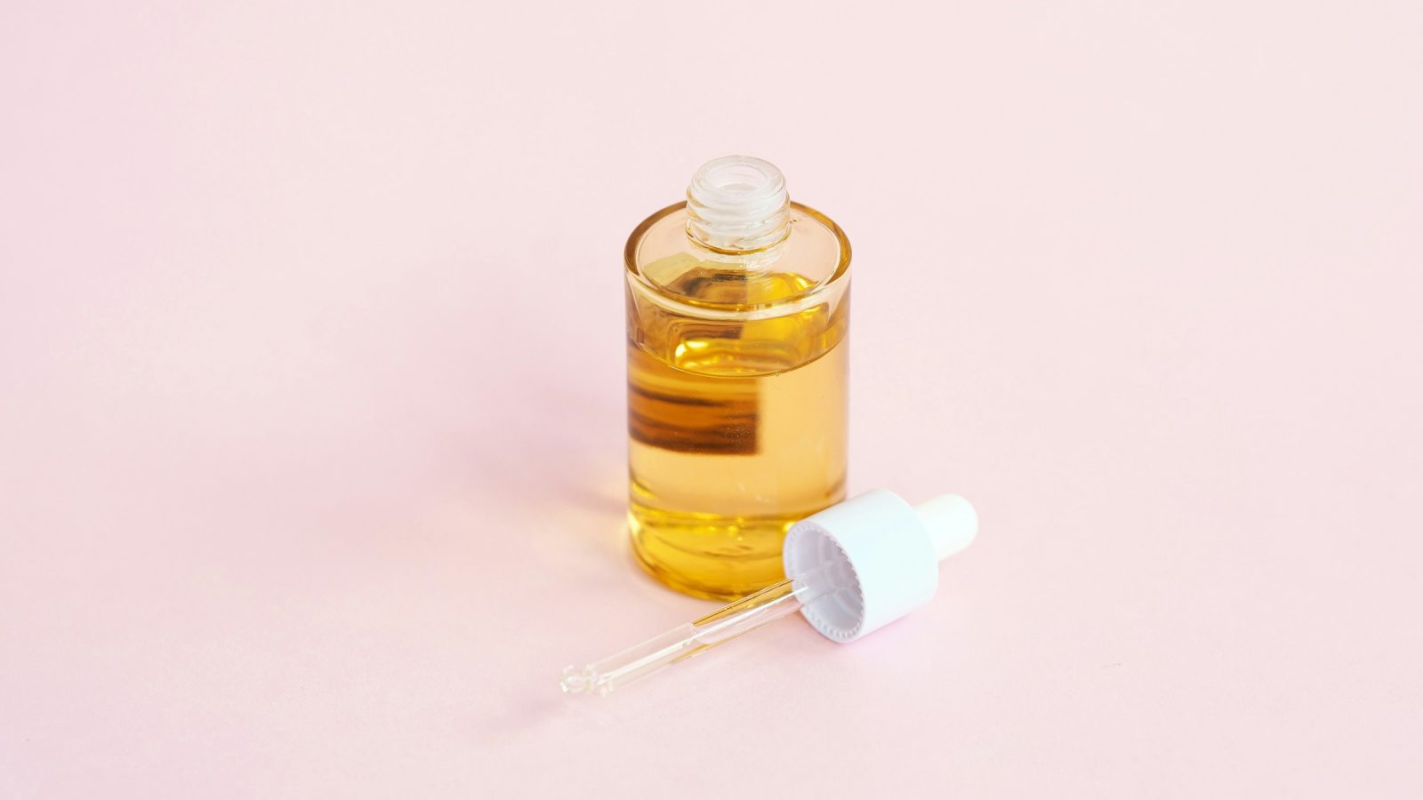 Glass bottle of golden hair oil with a dropper on a pink background