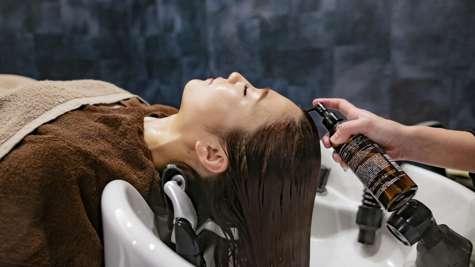 Stylist spraying a treatment onto a client's wet hair at the shampoo bowl